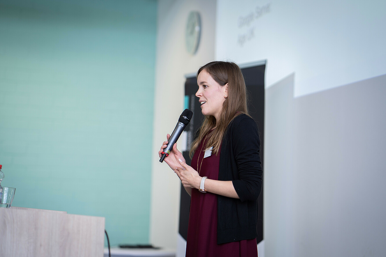 Eine Absolventin steht auf einem Podest im Vorlesungssaal, hält ein Mikrofon in der Hand und hält einen Vortrag.
