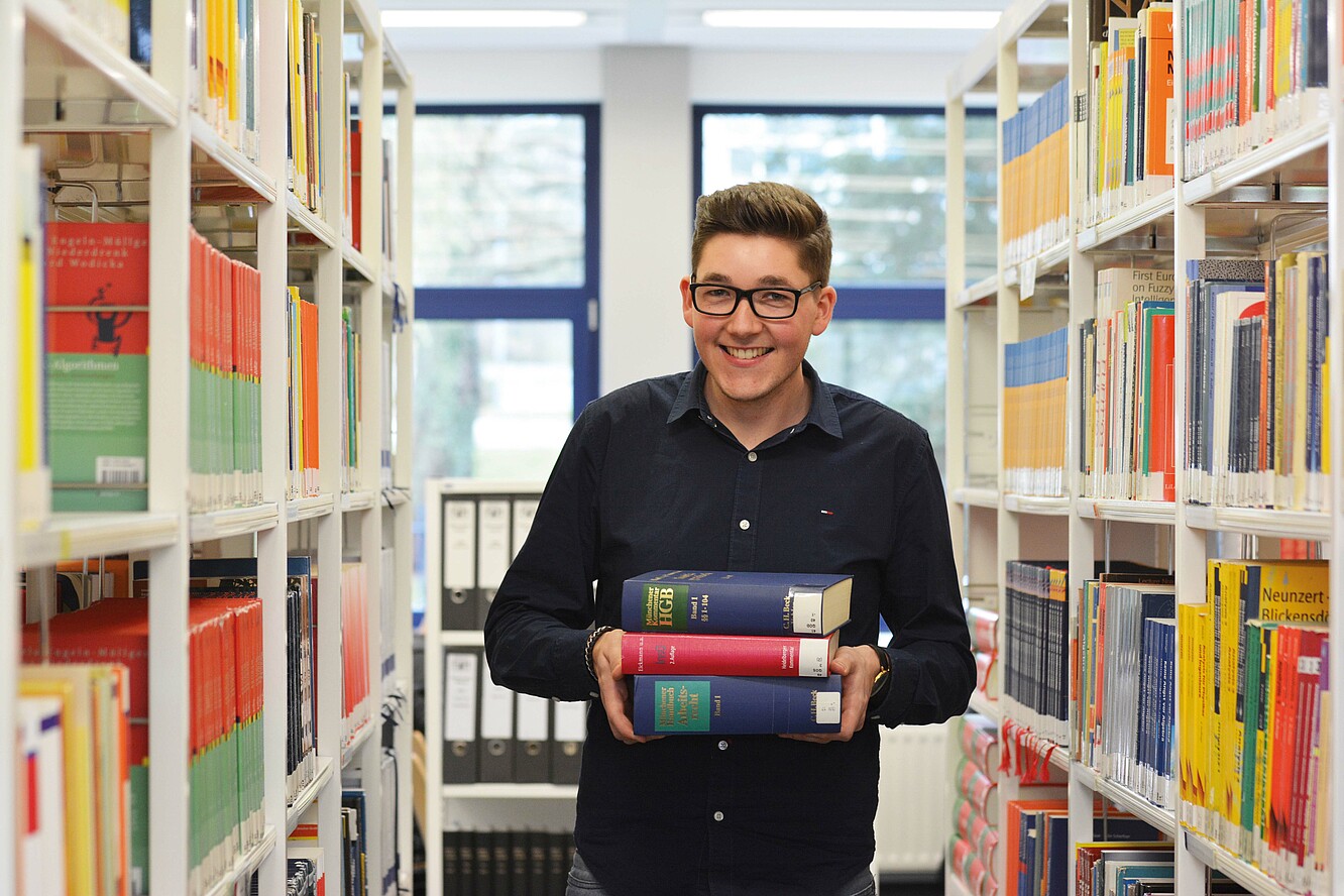 Ein Student steht zwischen zwei Bücherregalen, hat drei Bücher als Stapel in den Händen und lacht in die Kamera.