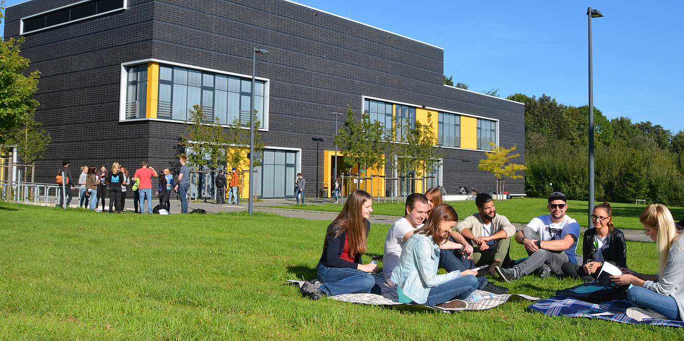 Studierende sitzen auf einer Wiese auf dem Campus Eupener Straße und unterhalten sich. Im Hintergrund sieht man das Hochschulgebäude D.