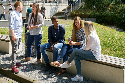 Drei Studierende sitzen auf einer Bank im Außenbereich der Mensa und unterhalten sich. Neben ihnen stehen zwei Studierende und unterhalten sich ebenfalls.